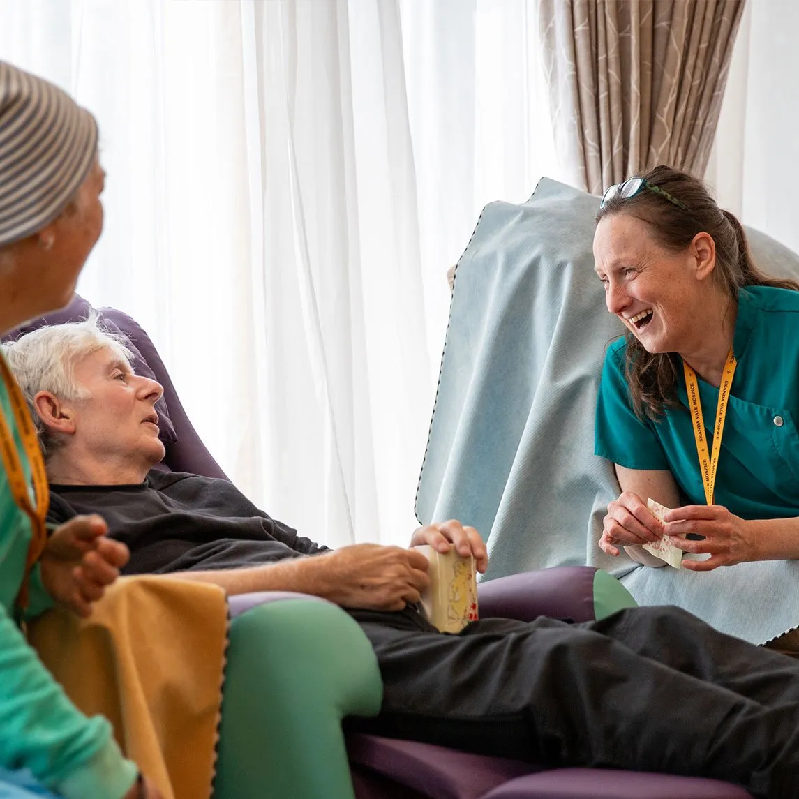 Nurse in teal scrubs laughing with two patients holding playing cards in reclining chairs beside a window.